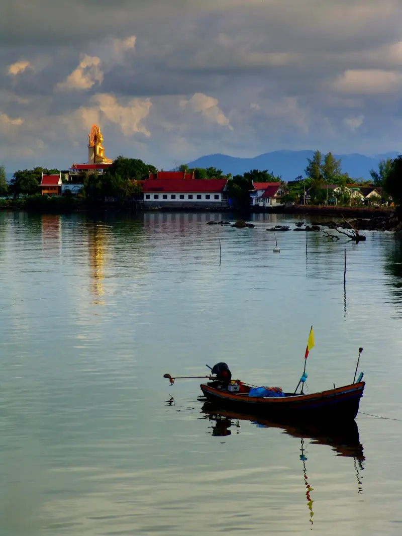 The Big Buddha on the shore of a bay with a fishing boat on the water in evening light