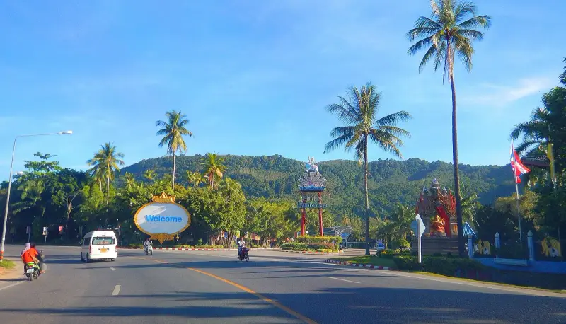 Дорога в Камале у въезда к Phuket FantaSea: пальмы, указатель Welcome и холм на заднем плане