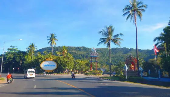 Дорога в Камале у въезда к Phuket FantaSea: пальмы, указатель Welcome и холм на заднем плане