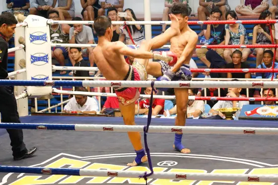 A Muay Thai bout: one fighter throws a knee, the other defends, spectators around the ring (shooting location not stated)