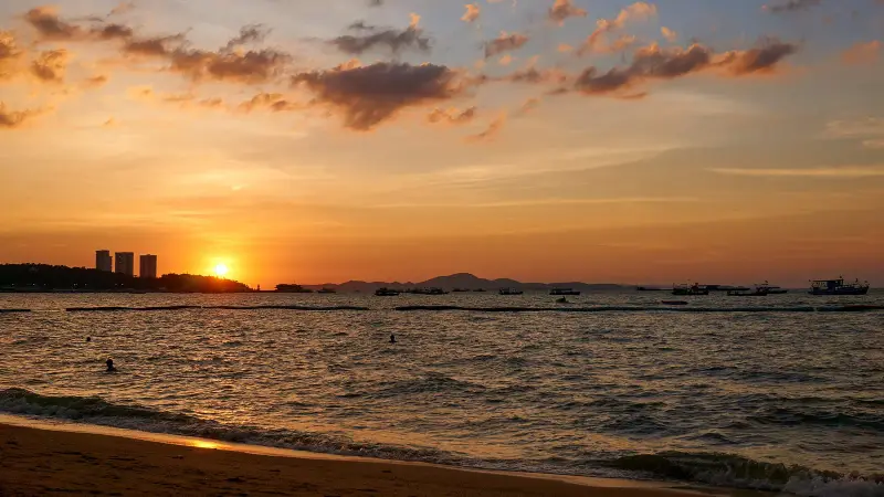 Sunset over Pattaya Bay seen from the beach: the sun sets behind a headland, boats stand on the water