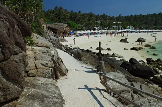 Path along the rocks to the Racha Yai beach with umbrellas and palms
